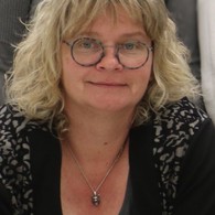 Eine blonde Frau mit Brille schaut neugierig in die Kamera.